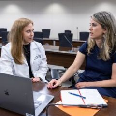 woman with long hair in white lab coat sits at laptop and talks with blonde woman in blue shirt