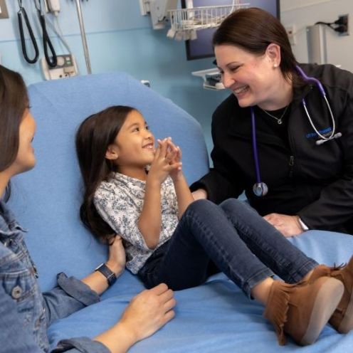 woman on left with jean jacket smiles at little girl on examination bed listening to medical worker woman with dark hair in dark scrubs with stethascope around neck