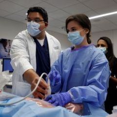 man in lab coat with surgical mask helps hold imaging scanner with woman in blue surgical scrubs and mask