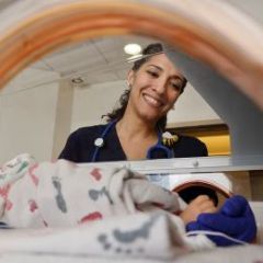 smiling dark haired woman in scrubs looks into incubator at baby in NICU