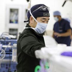 man wearing surgical mask and black scrubs works in hospital; man in blue scrubs in background