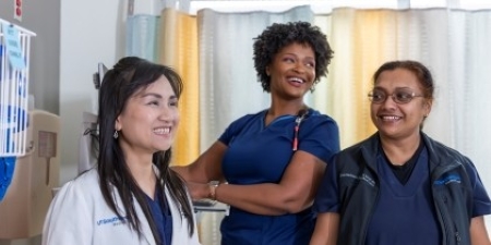 3 smiling women: dark hair in lab coat, tall woman with curly hair in blue scrubs, shorter woman with glasses in blue scrubs