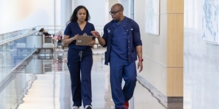 2 people in blue scrubs talk about notes on clipboard as they walk down hospital corridor