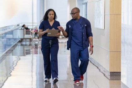 woman and man in blue scrubs walk down hospital corridor while discussing clipboard