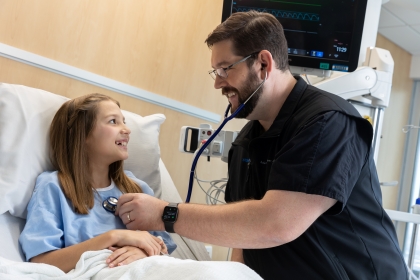 man in black scrubs smiles and uses stethascope on little girl in patient bed with hospital monitors in background