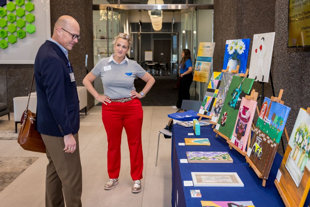 woman in red pants speaks to man in blue sweater looking at artwork on display table