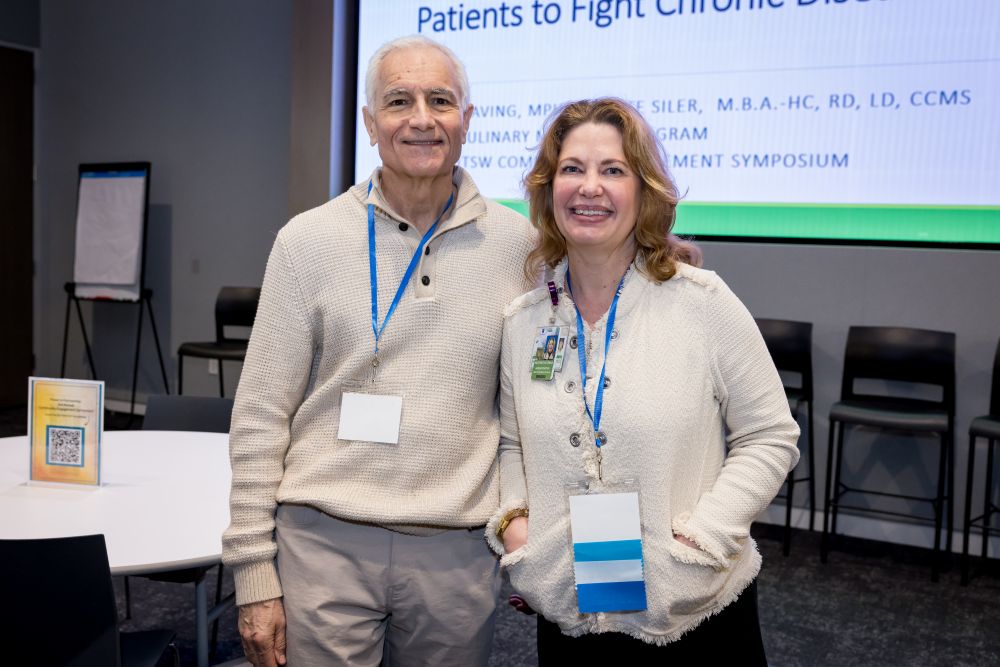 smiling white-haired man in sweater stands next to smiling blonde woman in front of presentation screen