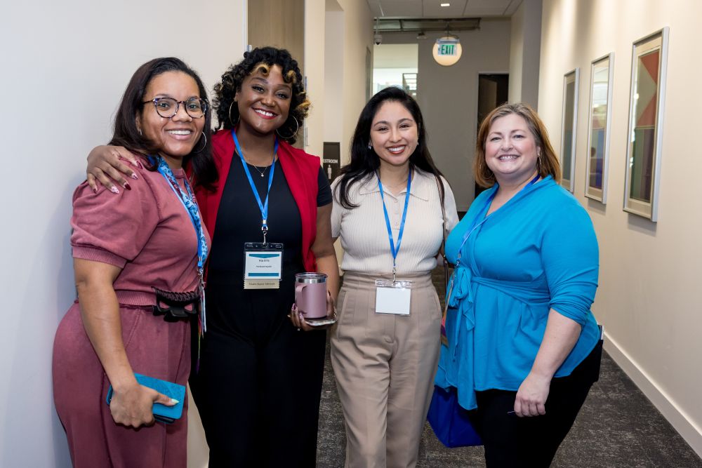 4 women stand together in hallway - from left, woman in dark pink outfit, woman in red blazer, woman with dark hair in cream sweater, blonde woman in bright blue top