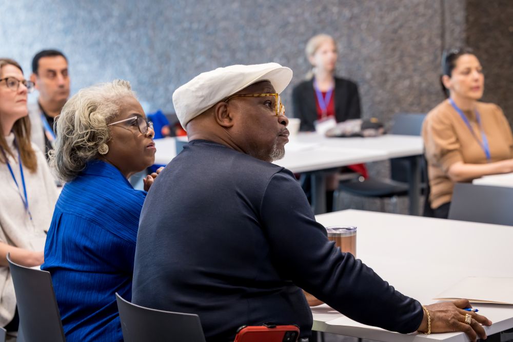 seated audience, including grey-haired woman in blue shirt and man with white cap and glasses, listens attentively