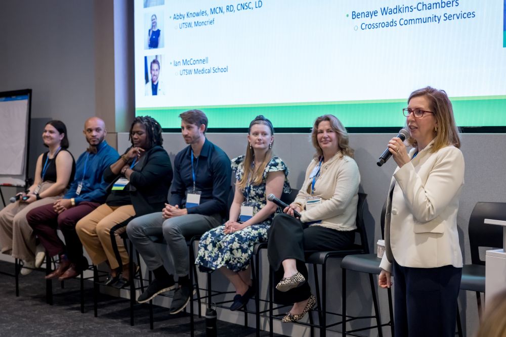 6 panelists seated across front of presentation screen as standing woman with microphone speaks to the right of photo