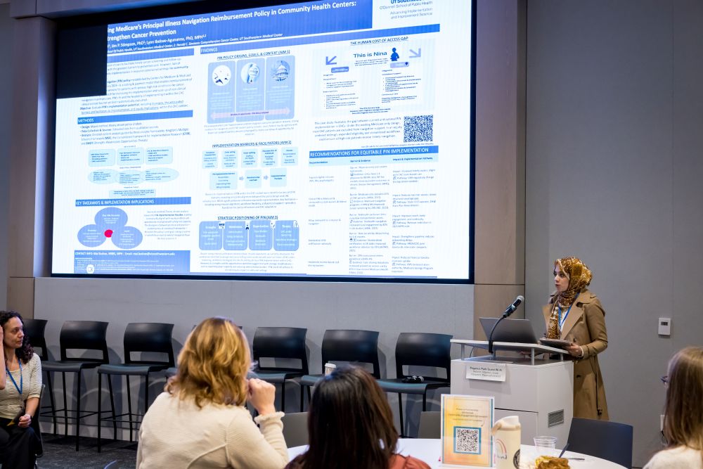 woman speaker at podium wearing gold patterned scarf and tan jacket shows slide on presentation screen behind her