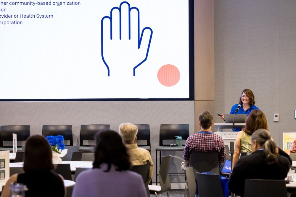 brunette woman speaks at podium to right as audience listens and looks at presentation slide with drawing of a hand