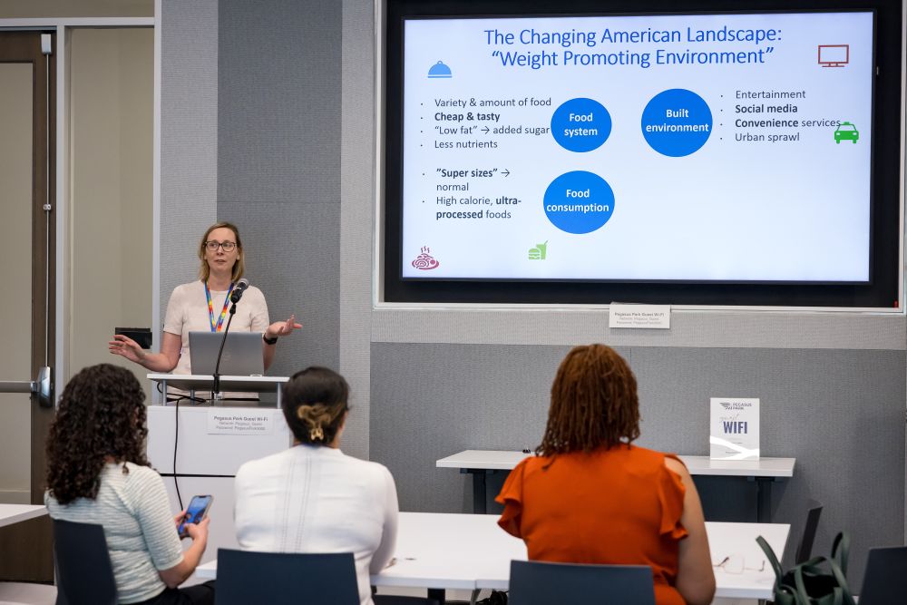 blonde woman speaks from podium left in front of presentation slide about The Changing American Landscape Weight Promoting Environment