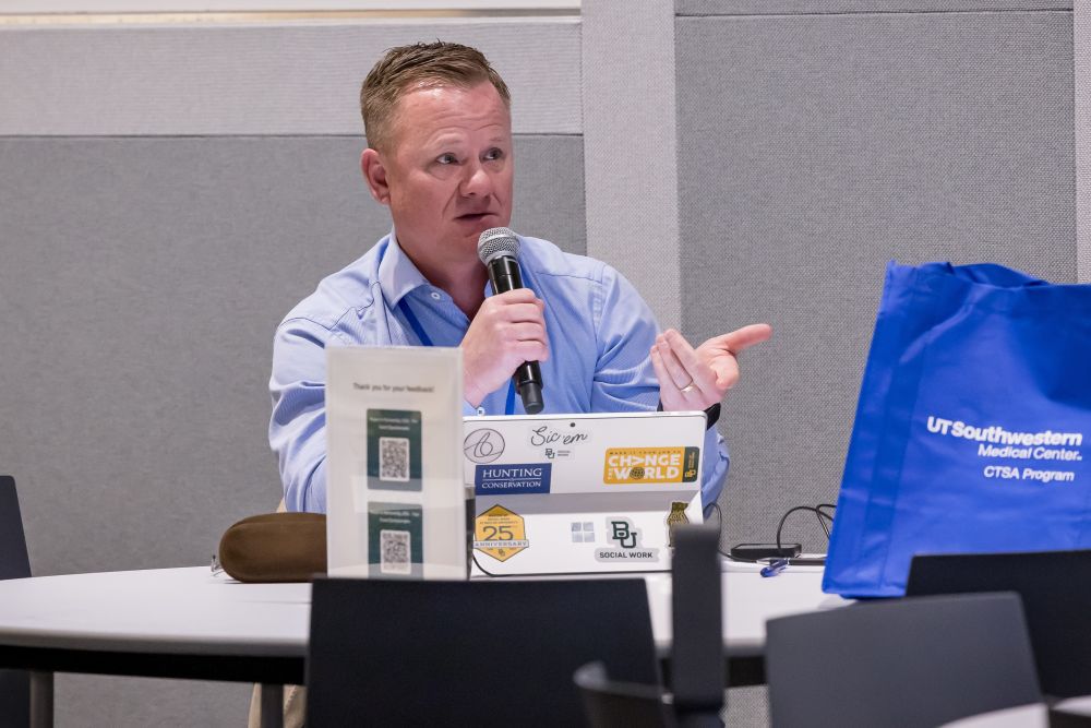 blonde man in blue oxford shirt seated at table asks questions during symposium