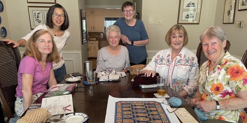 4 women seated at long dining table with needlepoint piece on table, 2 women stand behind the others
