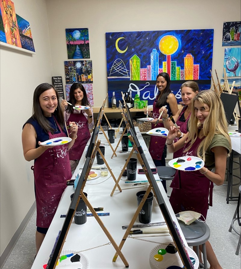 5 women wearing red aprons stand at painting easles holding paper plates with daubs of colorful pain