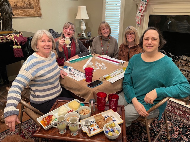 5 smiling women sit around mahjongg board in cozy room with cups and food plates in foreground