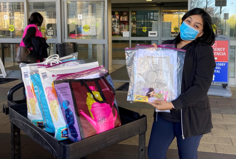 woman with dark hair wears surgical mask outside sliding doors, holds plastic bag containing art supplies next to cart with more bags