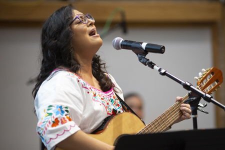 A woman in a colorfully embroidered shirt plays a guitar and sings into a microphone