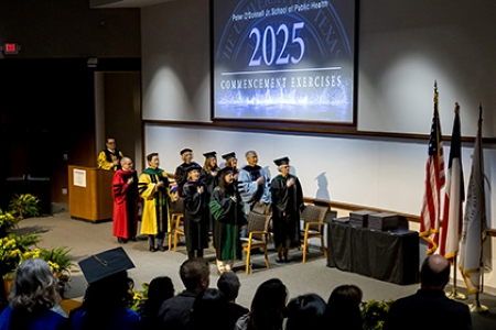 group of 10 faculty members on stage in cap and gown reciting pledge of allegiance to U.S. flag during O'Donnell School of Public Health 2025 commencement