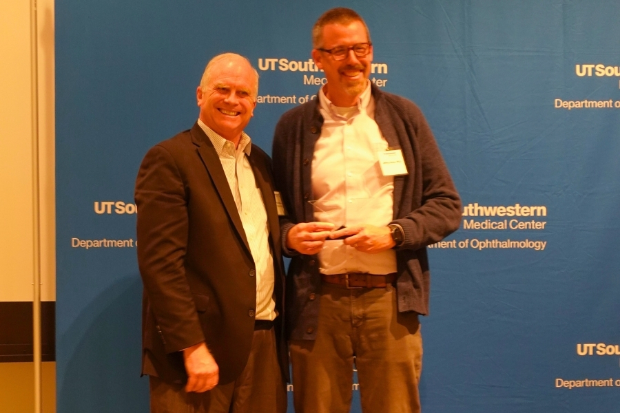 Two men standing in front of a blue Ophthalmology banner stand, one of them is holding an award he received