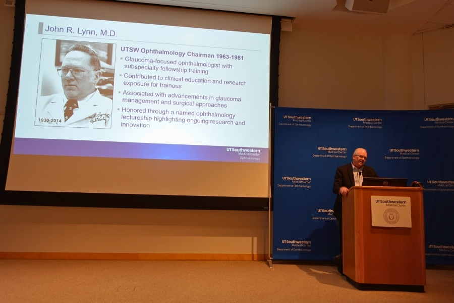 Man speaking at a podium in darkened auditorium next to a projected slide titled John R. Lynn, M.D.