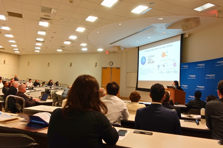 Seated conference attendees seated watching and listening to a female presenter at the podium explaining a slide call The Human Optic Nerve Cell Atlas