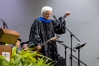 Man with white hair wearing academic robes holds a guitar and pumps his fist in the air.