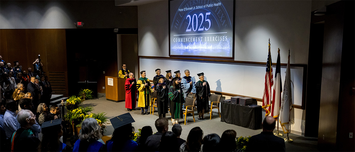 Speakers on stage at the Peter O'Donnell Jr. School of Public Health 2025, Commencement Exercises face the flags with right hands over their hearts.