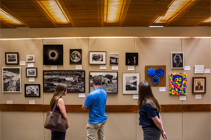 17 various art pieces on wall with 3 people looking at them