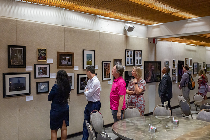 row of 7 men and women look at various artwork on gallery wall with round tables and chairs in foreground