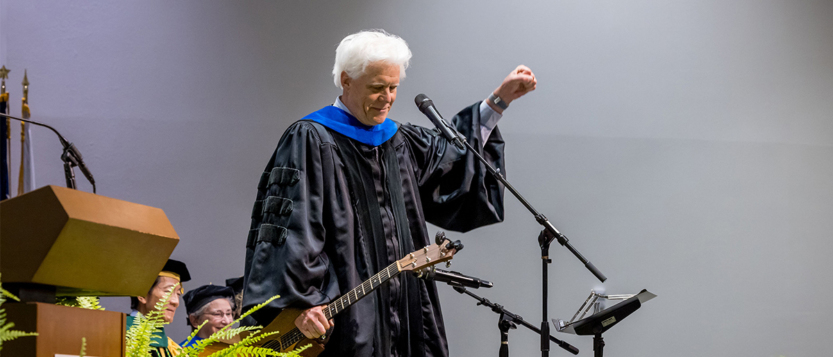 Man with white hair wearing academic robes holds a guitar and pumps his fist in the air.