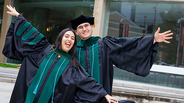 Two student wearing black and green robes, wave their arms as if spreading their wings.
