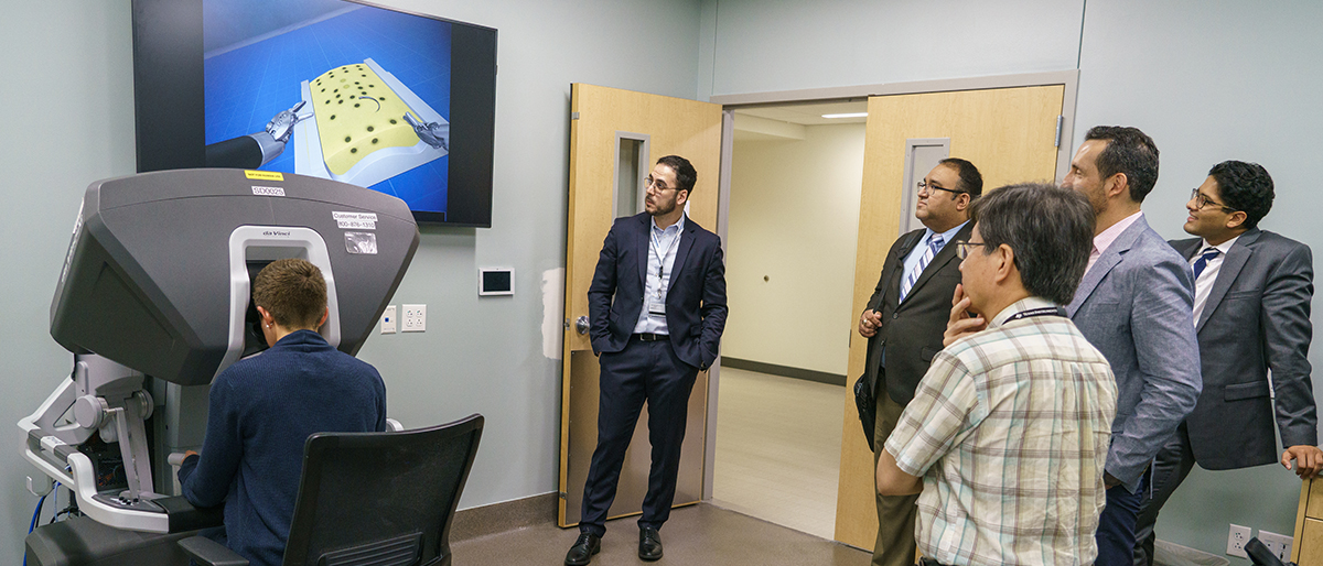 A seated person uses a simulation machine to make a suture and a group of men watch the the process on a large monitor.