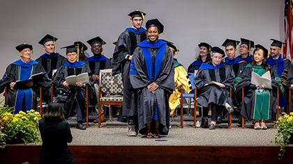 group of academics in cap and gown on stage behind podium; tall dark haired man with glasses stands behind smiling woman graduate to adjust her sash