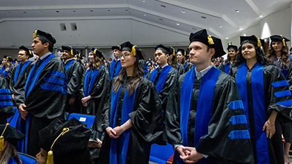 large group of graduates in cap and gown stand in auditorium