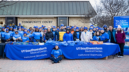 diverse group of employees dressed for cold weather outside  pose behind swag tables with UTSW blue covers and a building labeled Community Court in the background
