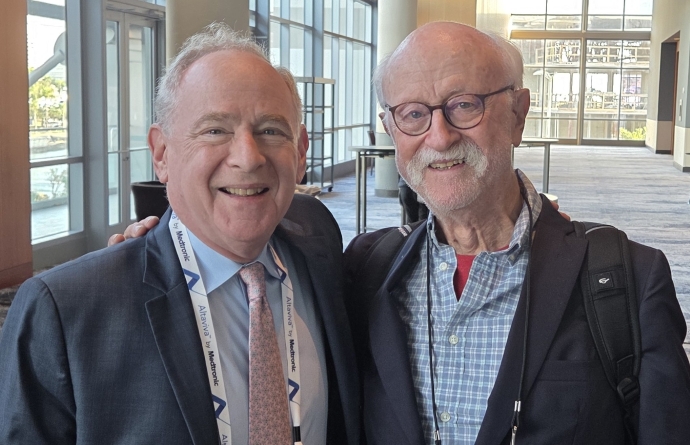 two older men stand side by side; man on left in blue suit, man on right with glasses and mustache