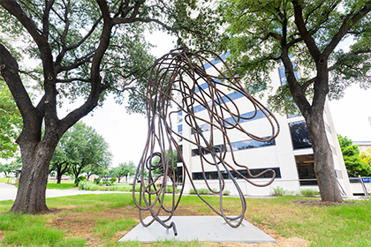 A large sculptureof twisted wire-like-iron stands between large trees in front of a building.