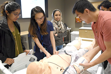 3 women students stand next to hospital bed as man demonstrates procedure on medical mannekin