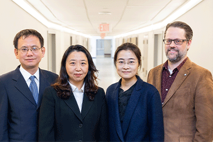 4 people in bright hallway: from left, man with dark hair and glasses in blue suit, 2 women with dark hair, tall man with glasses and beard in brown jacket