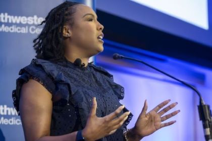 Woman with dark hair, wearing a blue dress, speaks into a microphone.