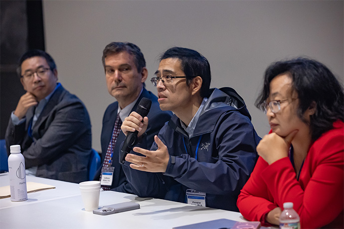 Three men and one woman seated at a table. One man is speaking emphatically into a microphone.