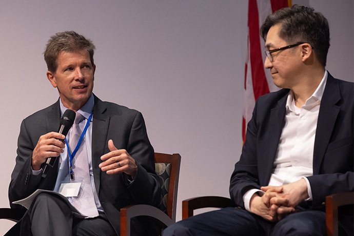 Man with short brown hair, wearing a gray suit, speaking into a microphone, and glancing at a man with dark hair and glasses wearing a dark suit seated next to him.