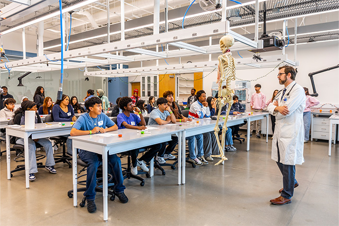 Students at tables observe a human skeleton.