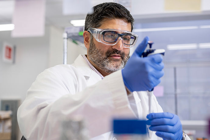 man with dark hair and grey beard wearing lab coat and safety glasses in research lab