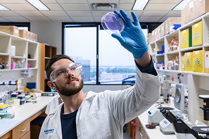 young man with beard wearing safety goggles in lab looks up at petrie dish