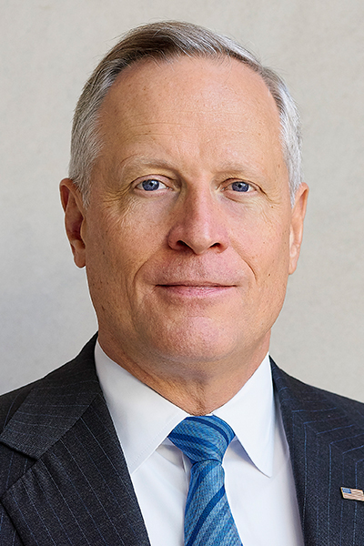 Man with short gray hair, wearing a dark suit, with a white shirt and blue striped tie.