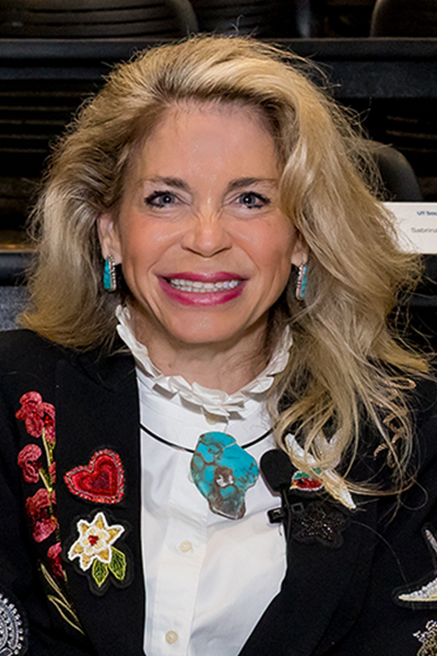 Smiling blond woman wearing a black jacket, seated at table.
