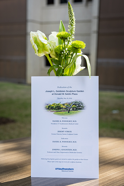 An invitation printed on white paper leaning against a vase of green and white flowers.